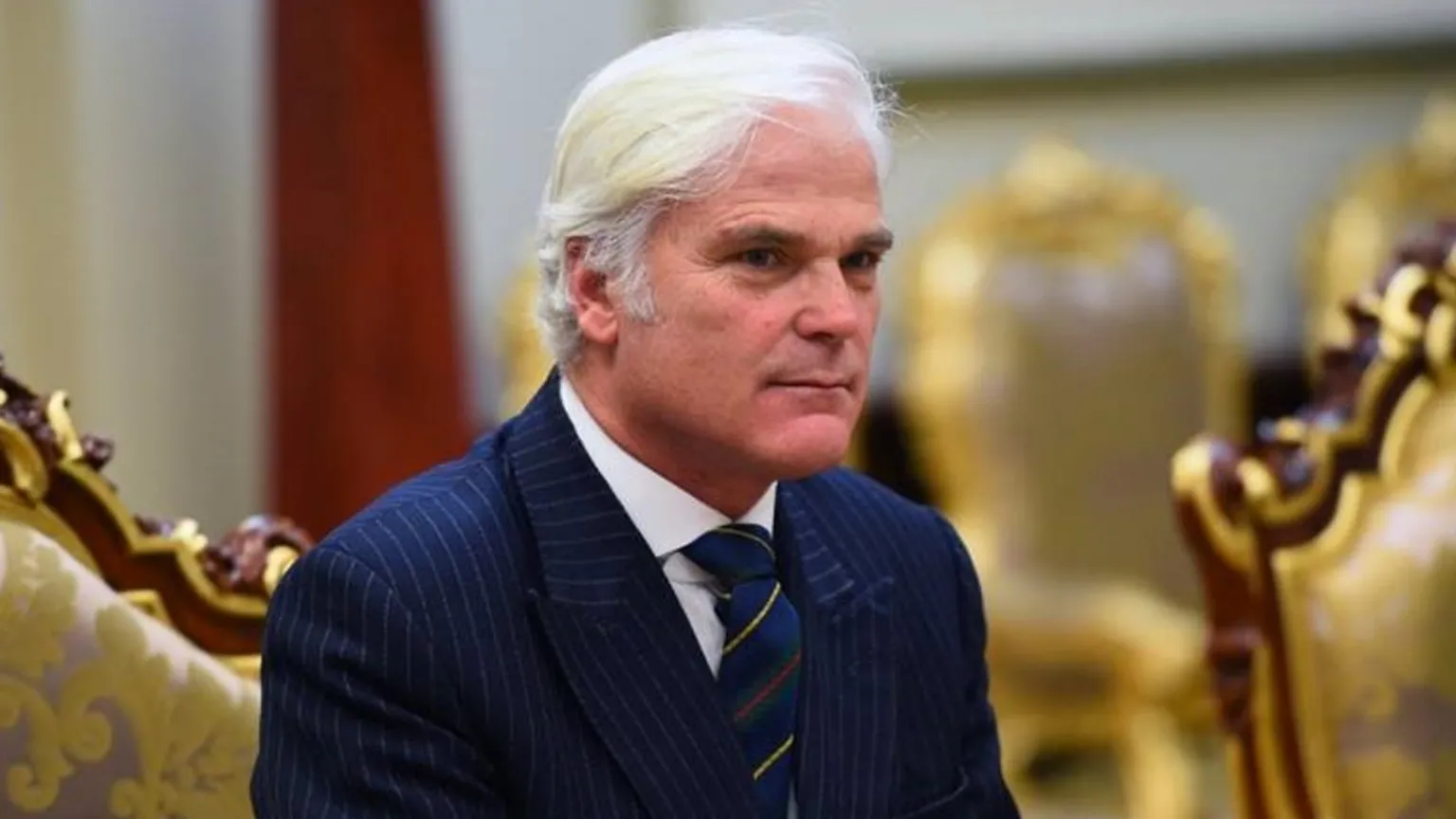 Getty Images Headshot of Sir Desmond sitting in a room with gold chairs. He has medium length white hair and wears a navy pinstripe suit with a white shirt and a navy and gold tie. 