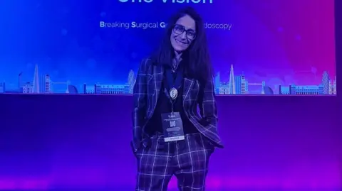 Rebecca Mallick A woman in a checked suit smiling at the camera. She is against a blue background. 
