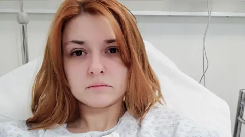 Eleni Moutesidi A woman sat in a hospital bed staring at the camera, she has ginger hair. 