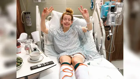 Lillie Bleasdale A woman in a hospital bed, she is making peace signs with both of her hands and looking towards the camera. 