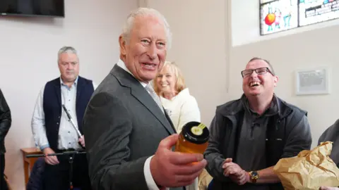 Reuters The King wears a dark-grey suit, white shirt and grey tie. He holds a clear jar of honey in his left hand. Her laughs as three people stand behind him. They are inside a building with white walls. A TV is over the King's right shoulder and a stain-glass window is over his left.