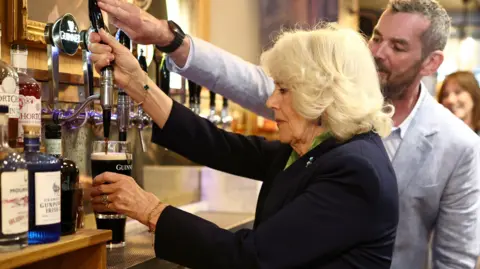 PA Media Queen Camilla wears a navy blazer and green shirt as she holds a partially full glass of black liquid, with a white head, in her left hand. She holds a black tap with her right hand. A man with short grey hair and a short brown beard helps her, his right hand is on the top of the tap. He wears a grey linen suit and a white shirt. 