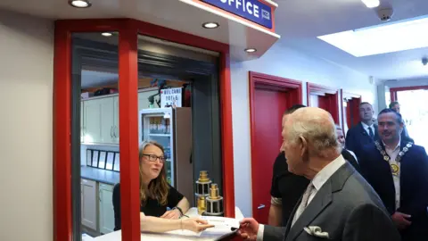 PA Media The King shakes hands with a woman with long brown hair, who wears a black t-shirt and glasses. She stands at the box office of a cinema. The king is wearing a grey suit, white shirt and grey tie. Behind the king are half a dozen people who watch on. 