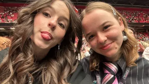 Nadia Wesseling Two girls close up to the camera, on the left she has wavy long brown hair and brown eyes. She is sticking her tongue out. On the right she has blond hair and disco ball earrings, she is smiling. Behind them are red stadium seats. 