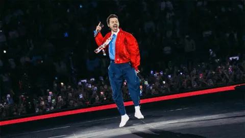 Anthony Pham/Getty Images Harry Styles is wearing navy trousers with a belt and a light blue shirt with a white collar and flower patterned tie.He has a red bomber jacket over the top. He is jumping on the stage, you can see fans watching from the crowd behind him.