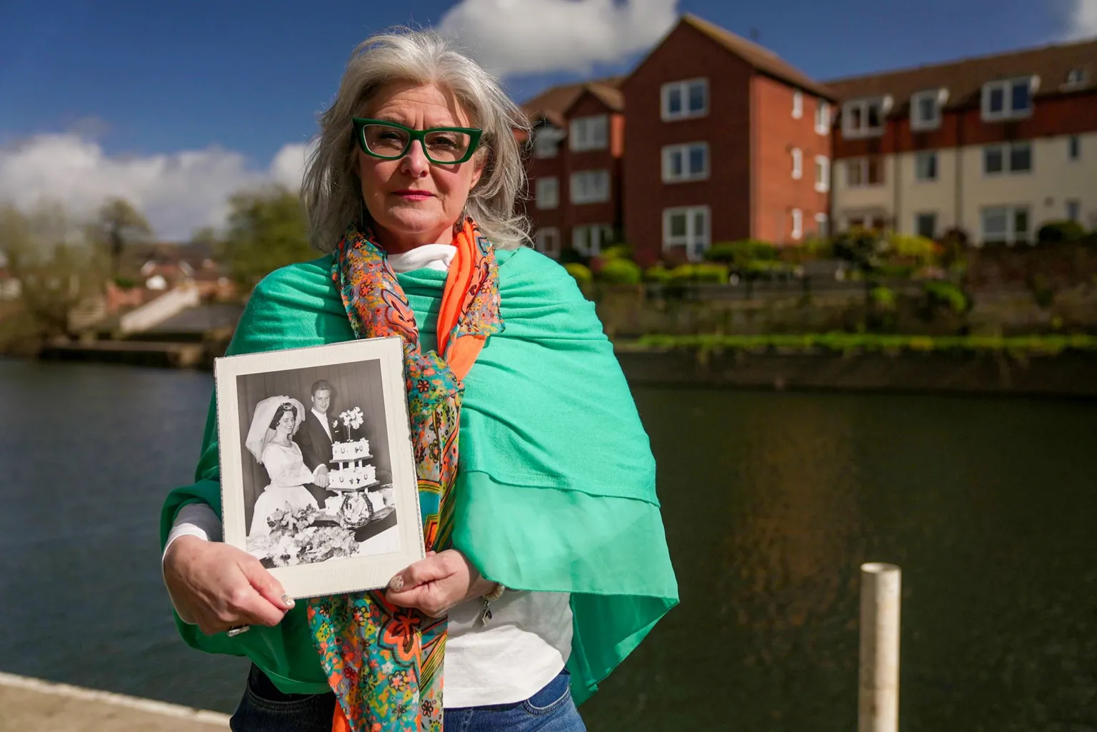 A woman in her fifties with flowing grey hair and strong, blue farmed spectacles stands by a canal in front of a small block of flats. She wears a bright green shawl, white shirt and orange scarf. She is holding a large black and white picture of her late parents on their wedding day.