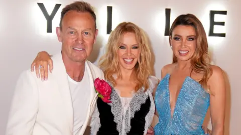 PA Media Kylie Minogue (centre) pictured with her arms around her sister Dannii and actor Jason Donovan
