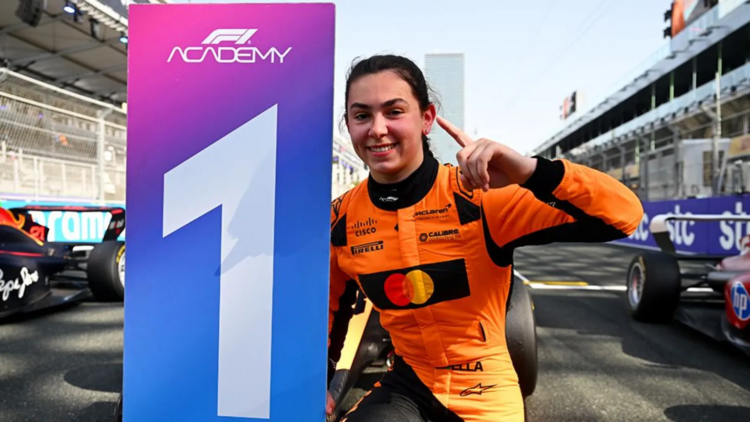 Getty Images Ella Lloyd, wearing an orange race suit with black sponsor logos, holding up a "one" sign with her fingers while crouching next to a "one" winners board. She is on the race track in front of a Formula 1 car.