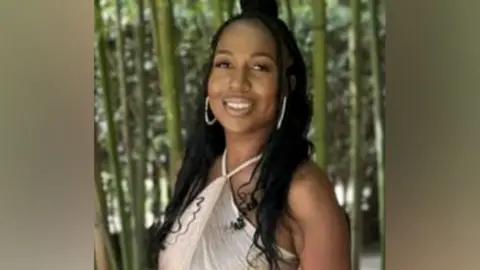 Family handout A young woman wearing gold jewellery smiles. She is standing in front of a forest.