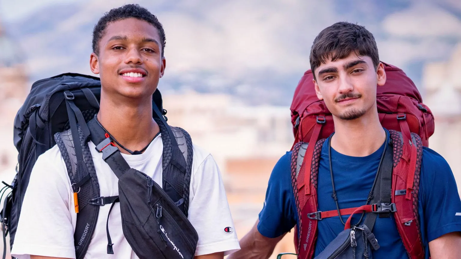BBC/Studio Lambert Jo and Kush, who are carrying large backpacks, are smiling at the camera at the start of Race Across The World in Italy. Jo is on the left and his wearing a white t-shirt with a black backpack, while Kush is wearing a blue t-shirt and has a red backpack. The background behind them is blurred but you can just about make out some blue sky and white clouds.