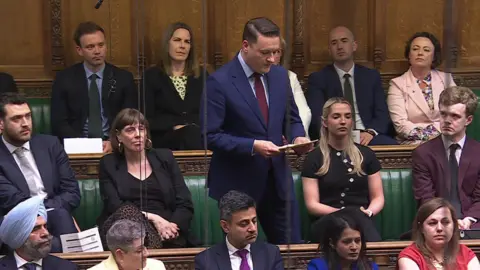 UK Parliament He is standing in the Commons chamber and wears a a pale blue shirt and maroon tie under a buttoned-up navy suit. He is reading from cards and is surrounded by Labour MPs who support his leadership bid.
