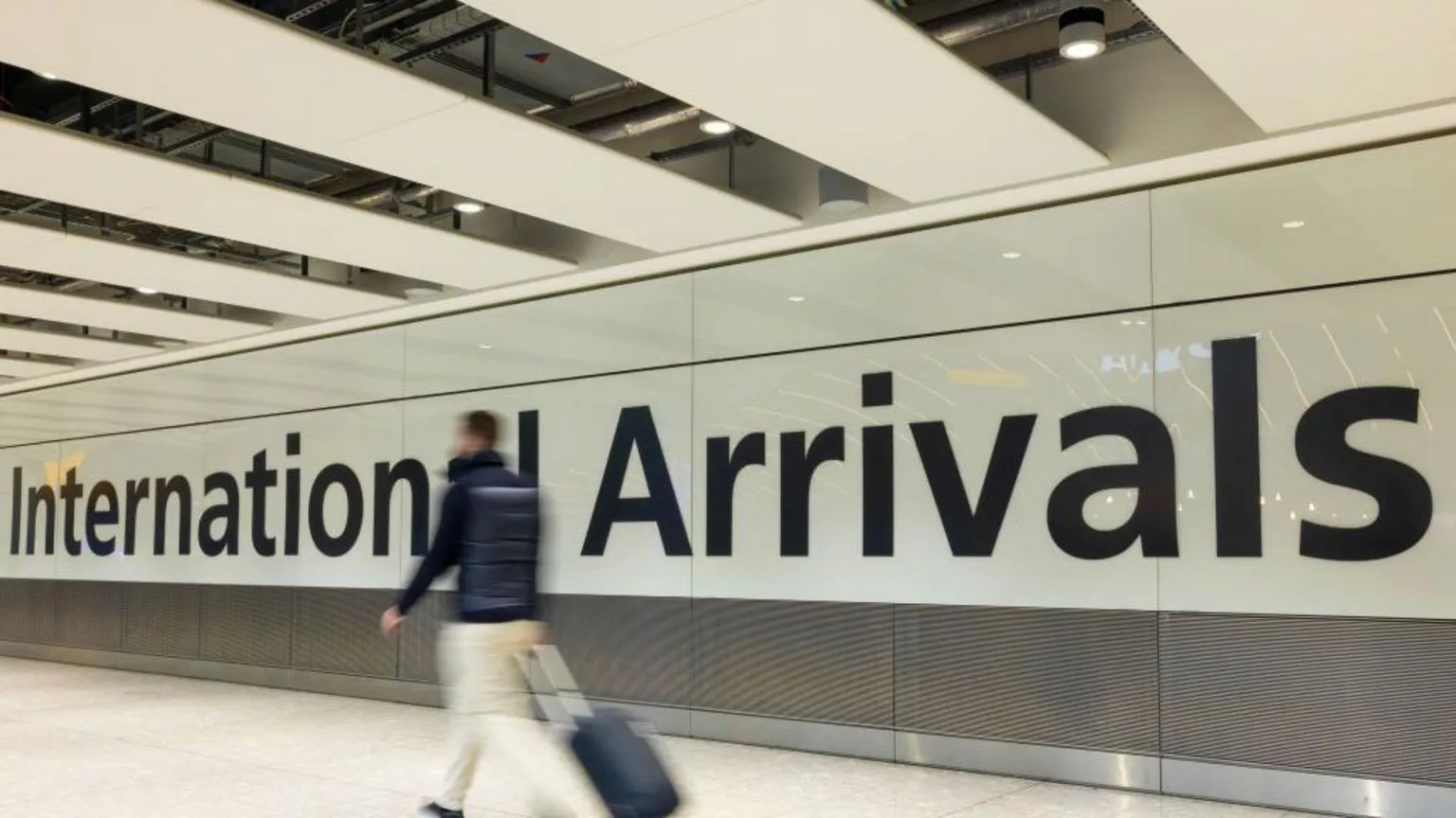Getty Images An airport corridor with the words 'International Arrivals' printed in large black letters onto a white wall. A man can be seen walking in front of the sign with a wheeled suitcase, beige trousers and a dark jacket, but he is slightly blurred to indicate the fact he is moving quickly. 